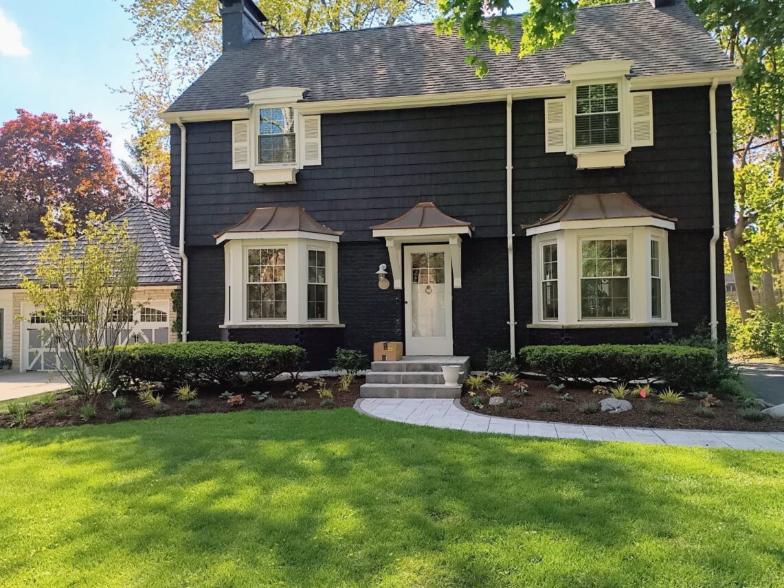 Two-story dark blue home with white trim featuring a stone paver walkway, manicured green lawn, and freshly mulched garden beds with low hedges and ornamental plants.