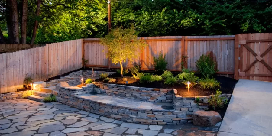 Wilmette backyard combining hardscape and plantings with stone patio, retaining wall, and uplighting.