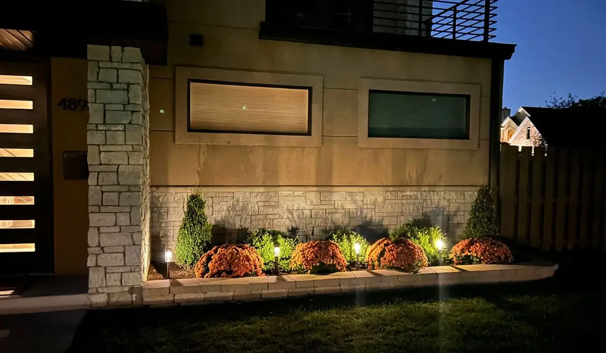Modern home landscape design featuring illuminated shrubs and flower beds with integrated all-season outdoor lighting system.