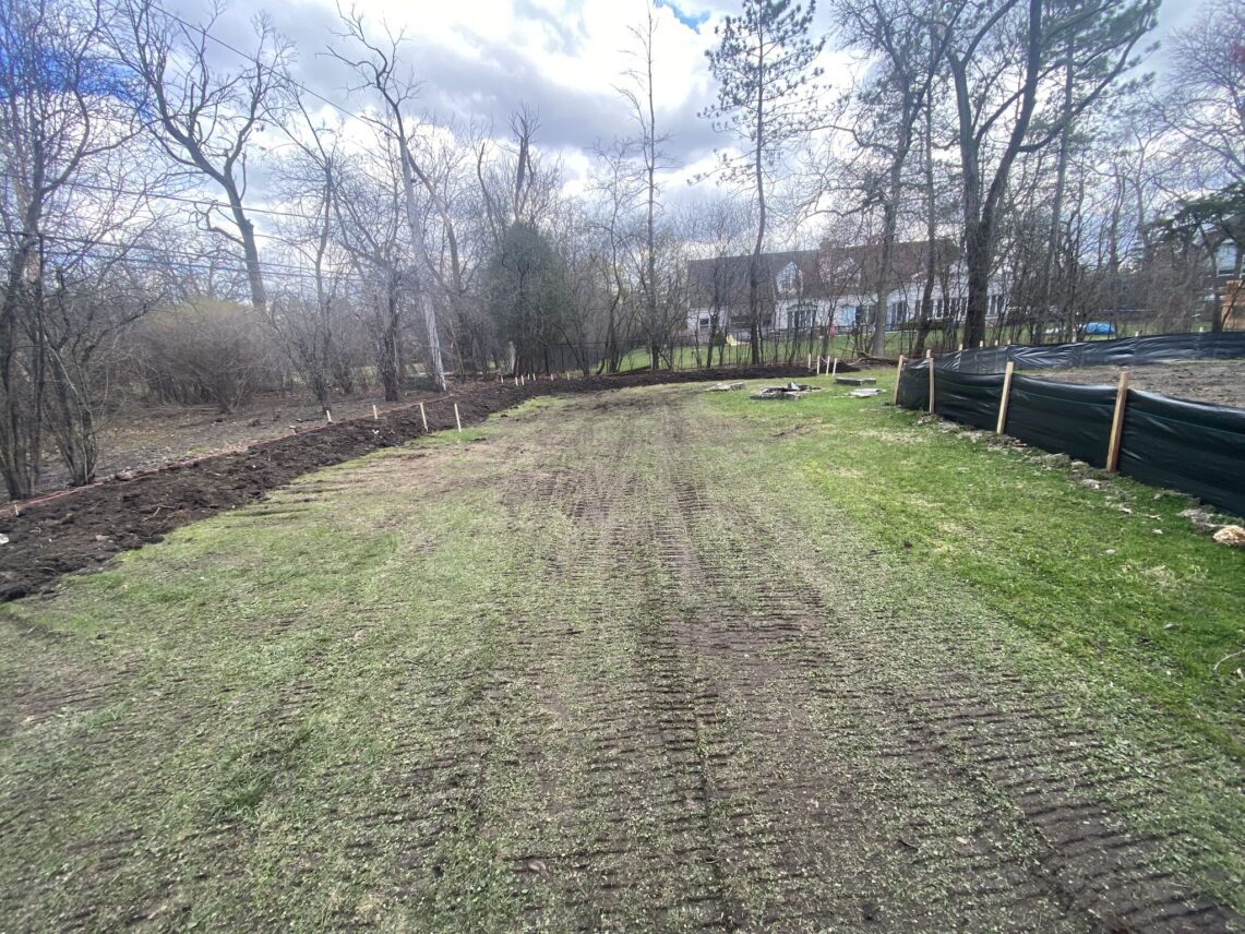 Large cleared yard area with visible tire tracks, a black silt control fence, and staked soil berms during a landscaping project in Northfield.