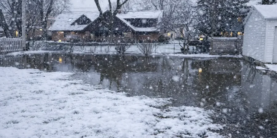 Snowy backyard with standing water after snowmelt showing winter drainage problems affecting lawn and foundation stability.